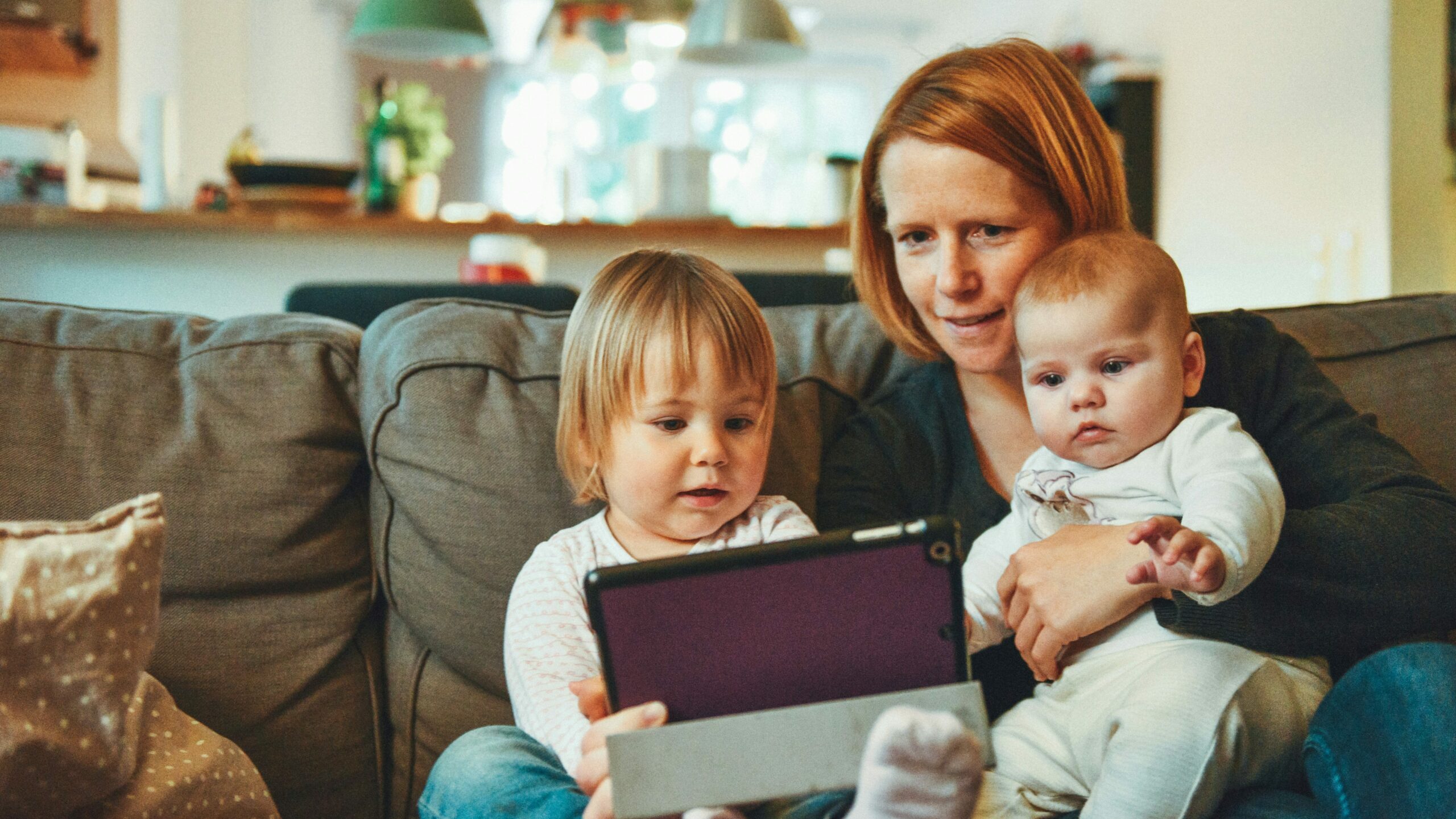 A mother comfortably sits with her children, including a toddler and a baby, showing a personal moment of family screen time. The trio is focused on a tablet, suggesting the early effects of kids' screen time on family bonding and child development.
