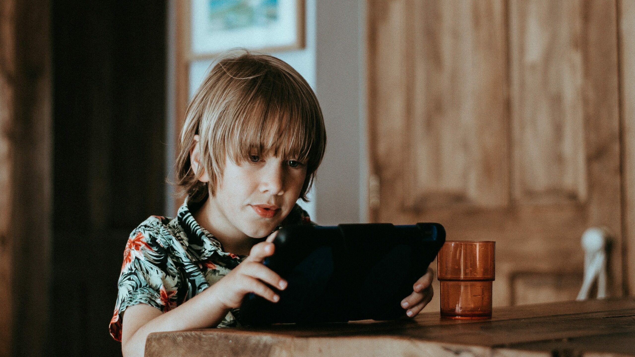 A young boy wearing a tropical print shirt, is absorbed in a handheld gaming device at a wooden table. This image highlights the effects of kids' screen time, portraying a child's deep concentration on digital entertainment, which is part of the ongoing discussion about technology's impact on youth.