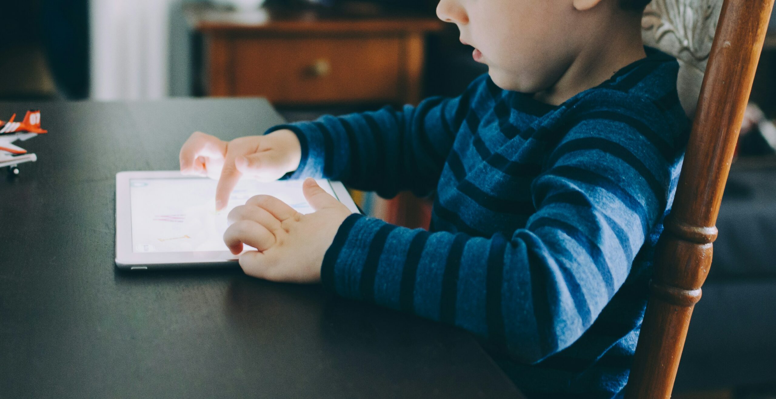 A young child, clad in a blue and dark striped sweater, is absorbed in a tablet, a scene that reflects the effects of kids' screen time. The child's fingers are poised on the touchscreen, illustrating the captivating nature of digital devices.