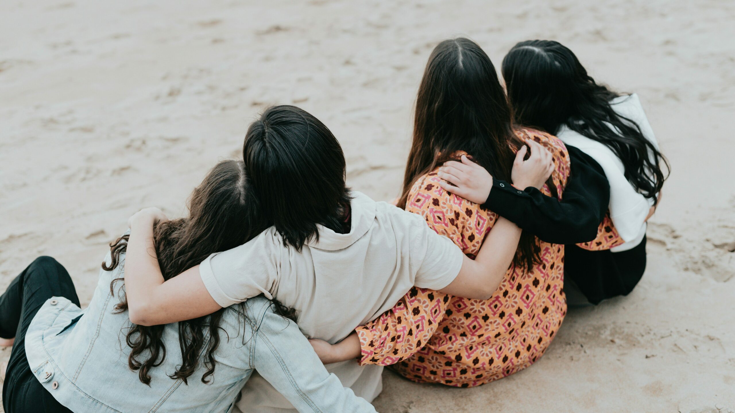 Four women sitting on the beach with their arms around each other, symbolizing solidarity and support. This image represents the community and assistance provided by domestic violence services.