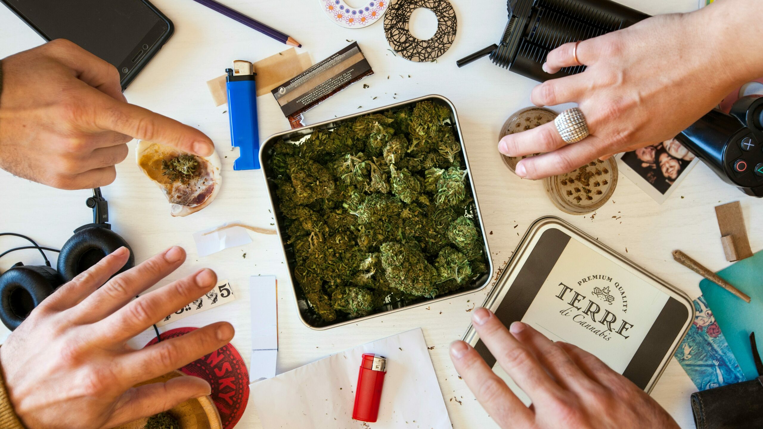 Top view of a table with various marijuana paraphernalia, including a tin of cannabis, a grinder, rolling papers, a lighter, headphones, and a smartphone, with multiple hands reaching in, highlighting 'marijuana use among youth.