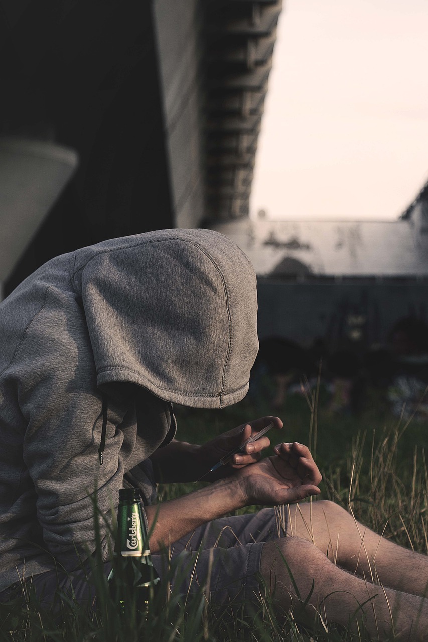 A person in a hooded sweatshirt sits in a grassy area under a bridge, holding a syringe as they prepare to inject a substance into their arm. A bottle of Carlsberg beer is on the ground beside them, suggesting summer substance use.