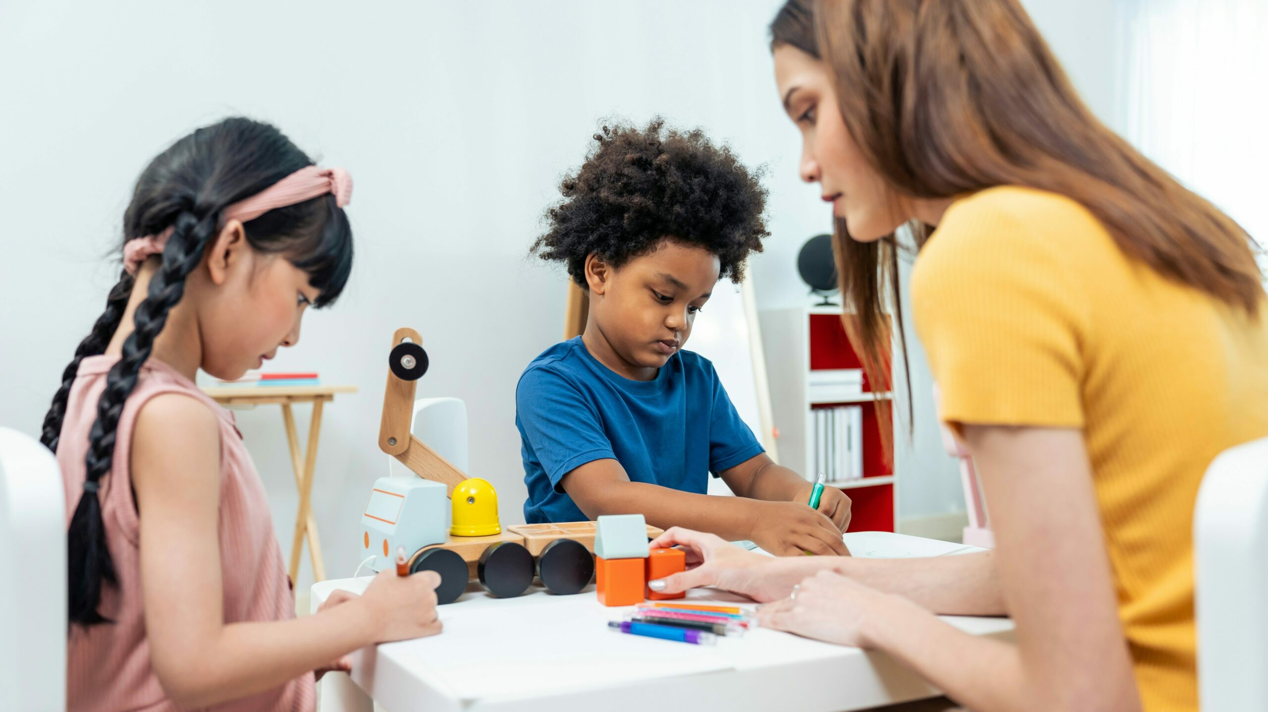 A caregiver provides HCBS prevocational support to three diverse children engaged in skill-building activities at a table, fostering inclusive early development.