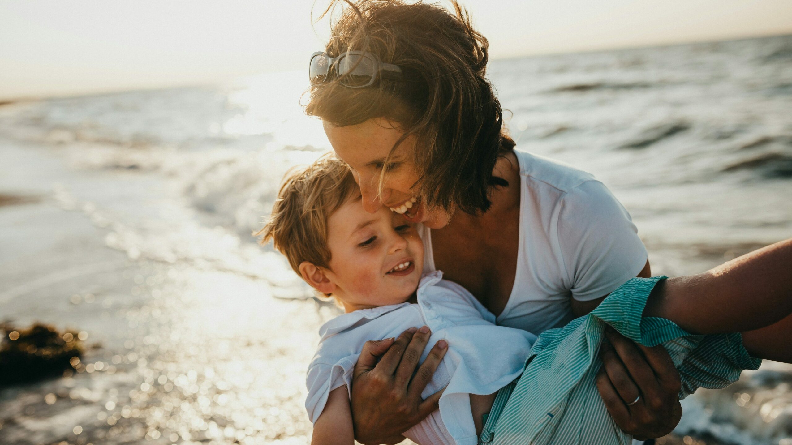 Mother holding her child on a beach during sunset, both smiling and enjoying a moment of togetherness, representing the nurturing environment of Children's hHome and Community-Based Services (HCBS).